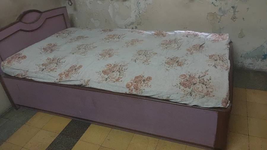 Lilac single bed with a decorative headboard and a rose-patterned sheet, against a wall with peeling paint.