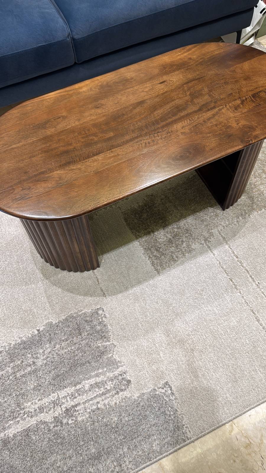 Oval imported coffee table with dark brown wood top and unique fluted base, near a blue sofa on a grey rug.